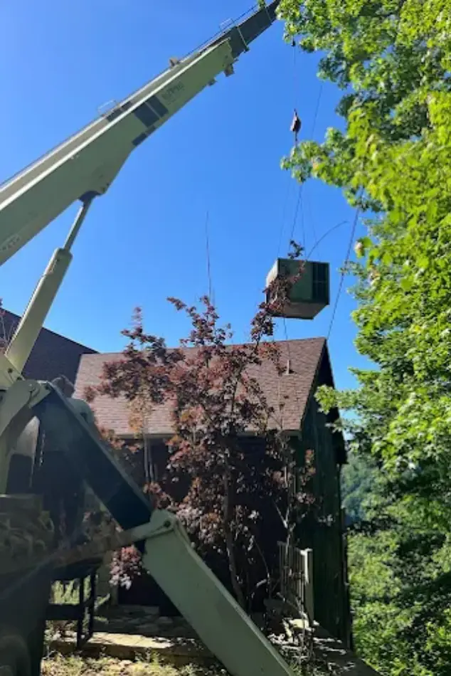 A crane lifts an air conditioning unit toward a house, surrounded by green trees and a clear blue sky.