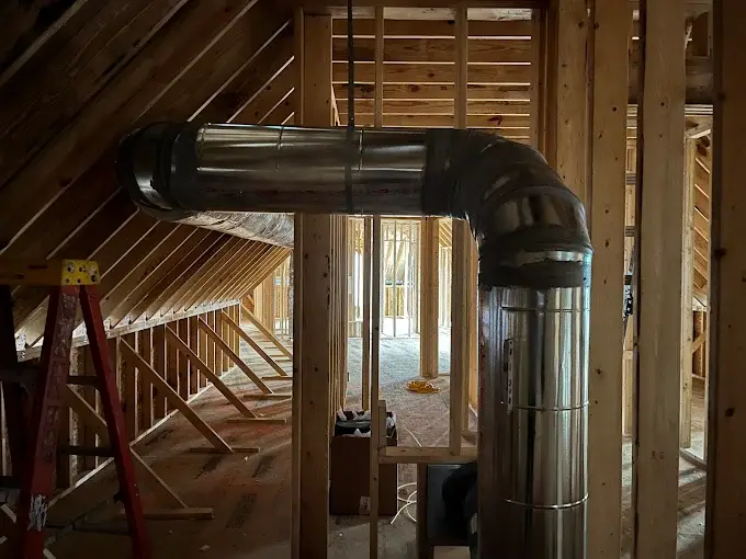A metal duct curves through a wooden-framed attic space, with construction tools and materials visible in the background