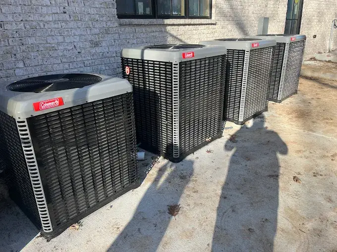 Four black outdoor air conditioning units are lined up against a brick wall, casting shadows on the ground in bright daylight